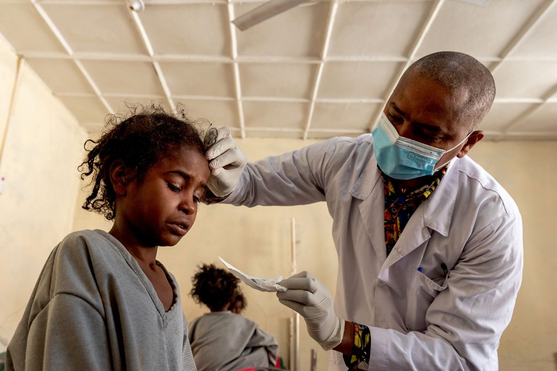 Ein Mädchen schaut mit geschlossenen Augen nach unten, ein Mann hat eine Hand auf den Kopf des Kindes gelegt, in der anderen Hand hält er einen Verband.