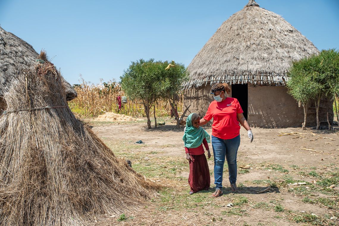 Eine Frau in Jeans und T-Shirt mit CBM-Logoe geht neben einem etwa neunjährigen Mädchen mit Kopftuch zwischen Rundhütten entlang.
