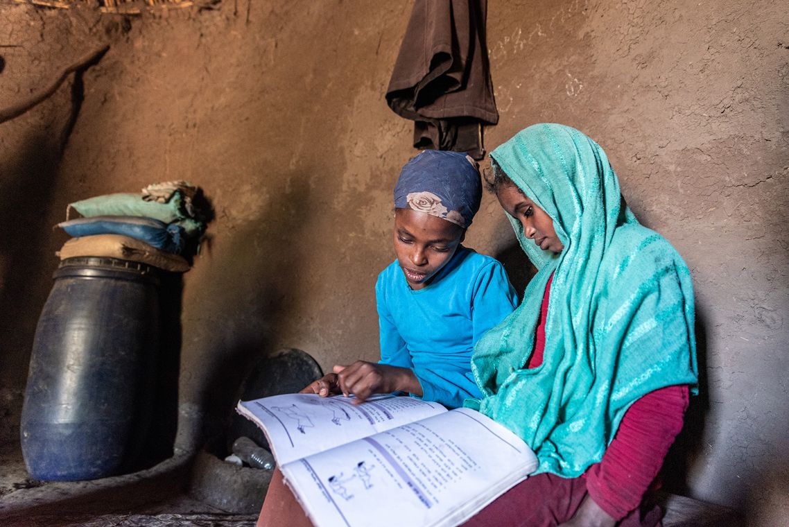 Ein etwa neunjähriges und ein etwas größeres Mädchen sitzen nebeneinander in einer Lehmhütte. Das größere Mädchen liest aus einem Heft vor.