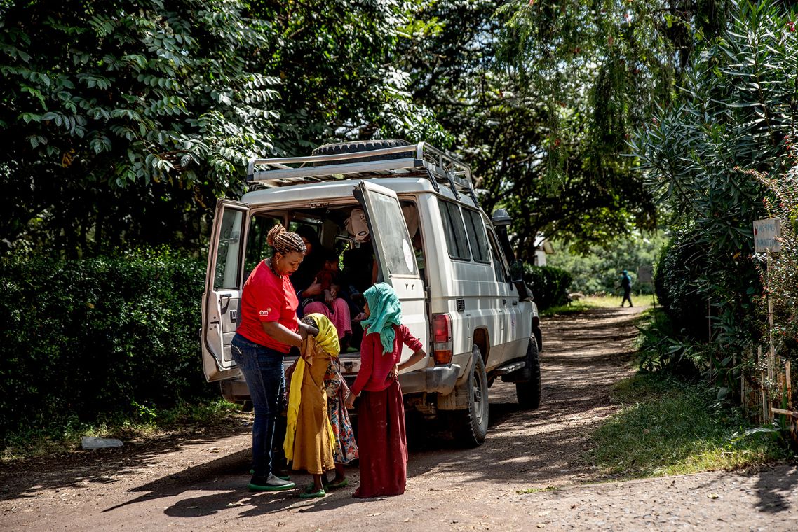 Eine Frau und zwei kleine Mädchen stehen hinter einem Kleinbus vor dessen geöffenten Türen.