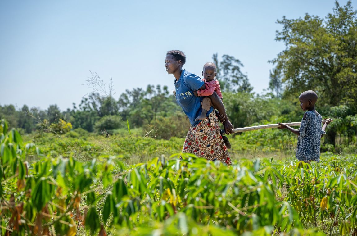 Eine Frau mit einem Kleinkind auf dem Rücken läuft mit einem Stock über ein Feld. Am anderen Ende des Stocks hält sich ein Kind fest.