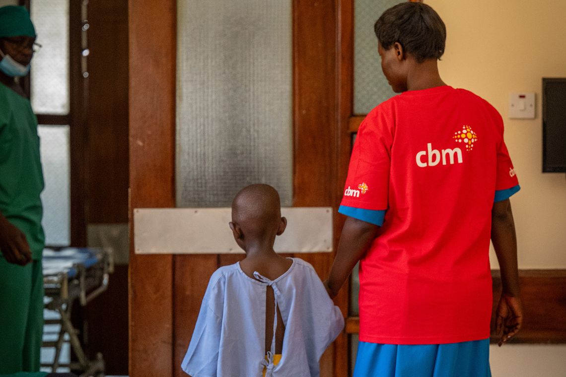 Eine Frau in einem roten T-Shirt hält die Hand eines sechsjährigen Mädchens, das Krankenhauskleidung trägt.
