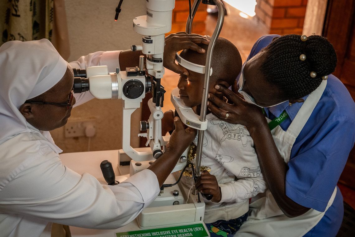 Eine Frau untersucht ein Kind mit einer ophalmologischen Spaltlampe. Eine andere Frau sitzt hinter dem Kind und hält sanft den Kopf des Kindes in Position. 