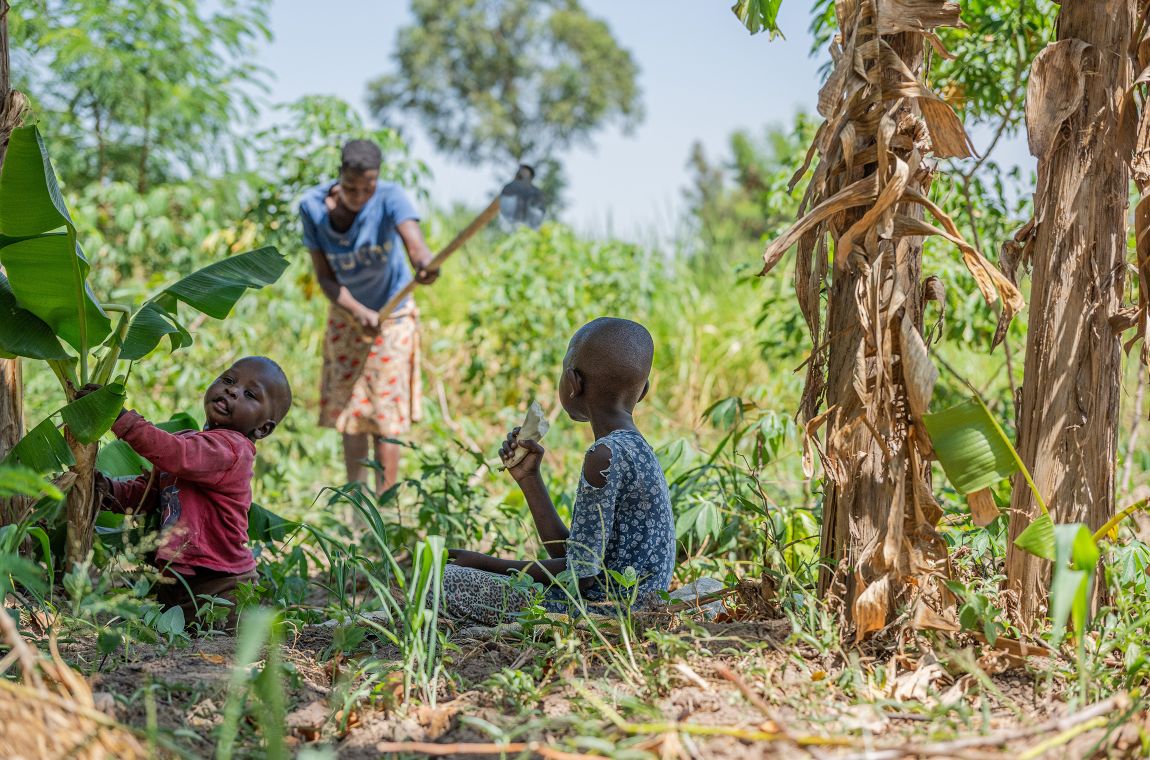 Eine Frau arbeitet mit einer Hacke auf einem Feld. Am Feldrand sitzen zwei kleine Kinder.