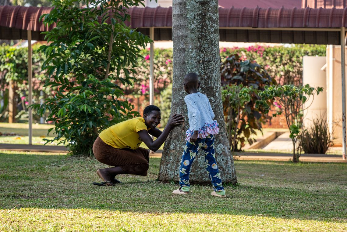 Eine Frau sitzt gebückt an einem Baum. Ein sechsjähriges Kind geht auf die gebückte Frau zu. 