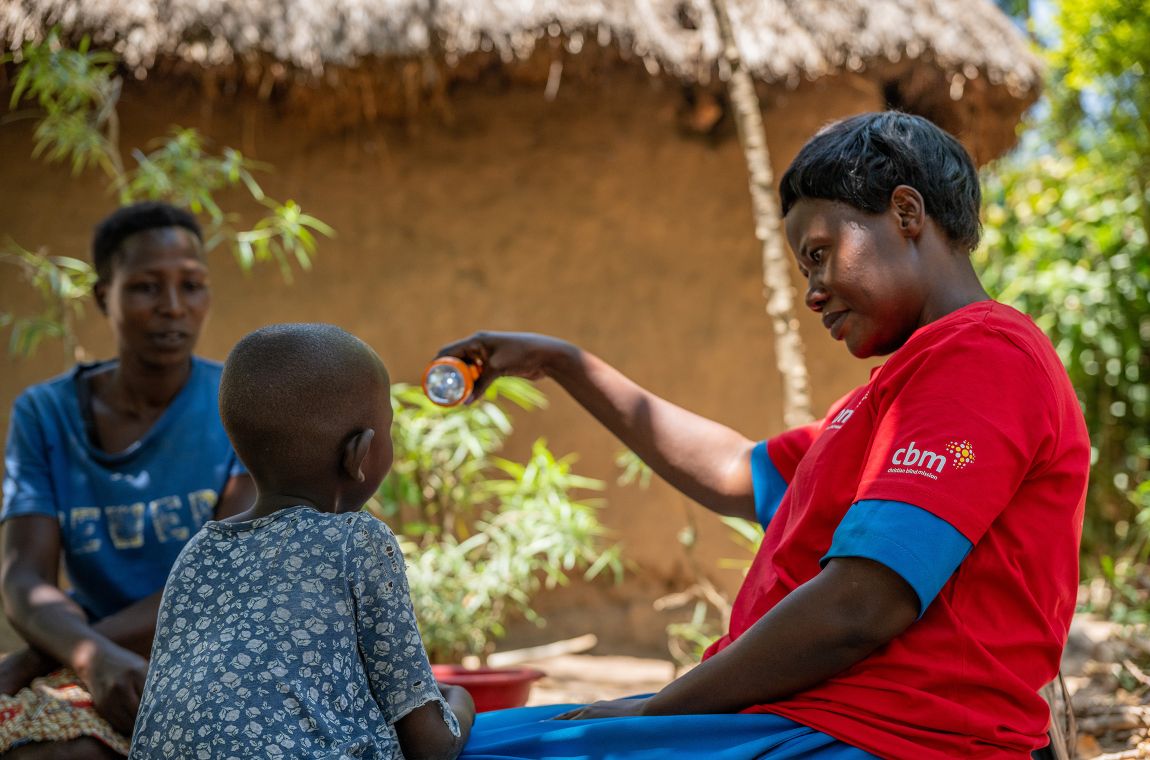 Zwei Frauen und ein kleines Mädchen sitzen auf einer Decke. Eine Frau leuchtet dem Mädchen mit einer Lampe in die  Augen.