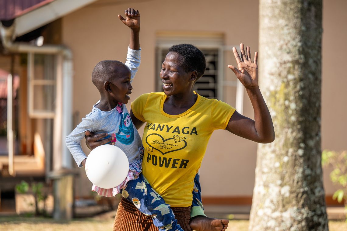 Eine Frau hat ein Kind auf dem Arm. Beide lachen und heben jeweils eine Hand in die Luft. 