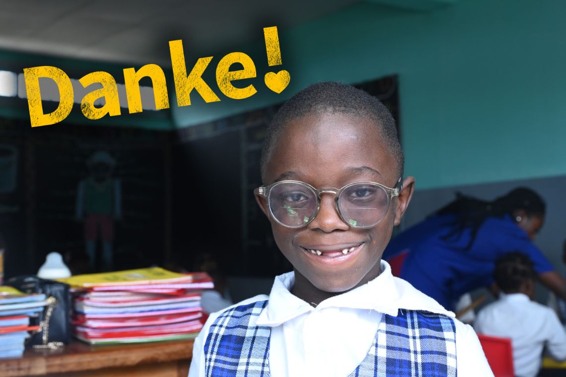 Ein etwa fünfjähriger Junge mit Zahnlücke trägt eine große Brille. Er trägt ein weißes Hemd und eine karierte, blau-weiße Weste und steht lächelnd in einem Klassenzimmer.