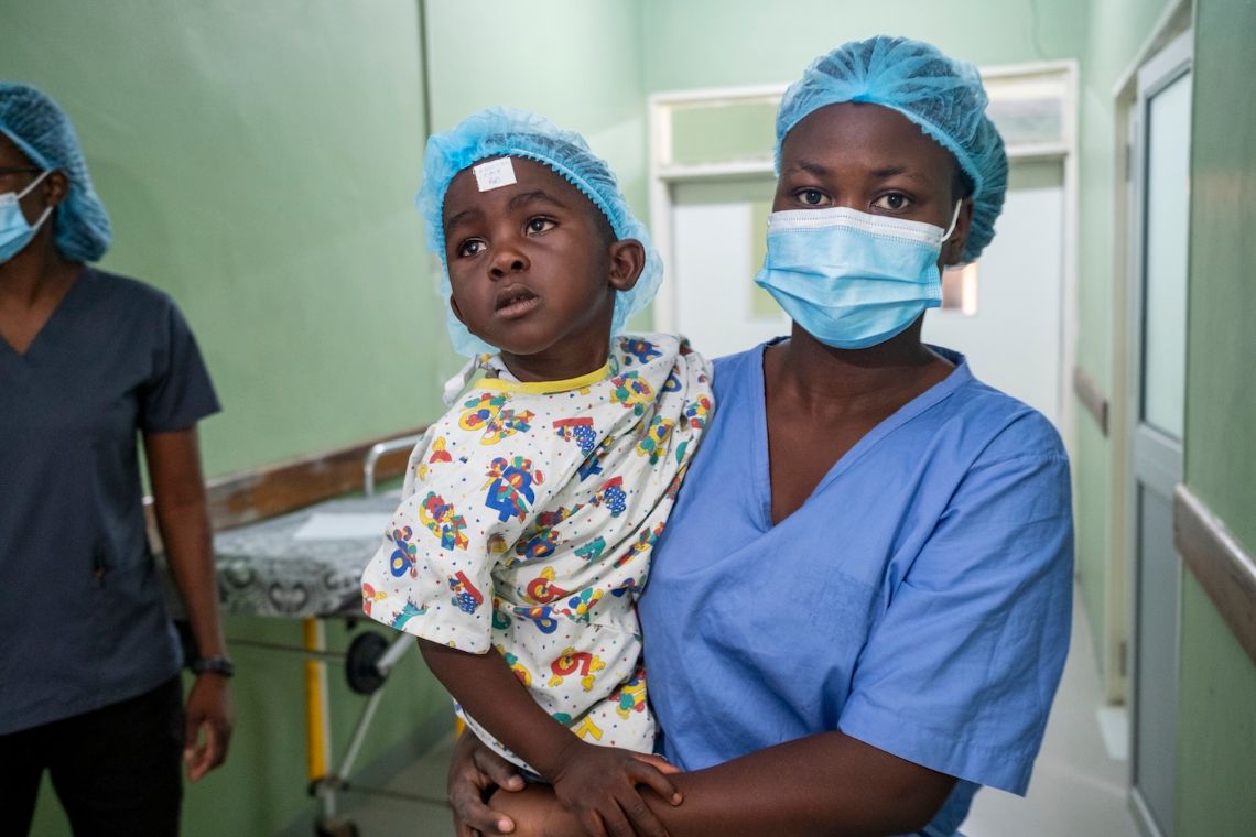 Ein etwa dreijähriger Junge mit OP-Haube wird von einer Frau in OP-Kleidung durch einen Flur getragen.