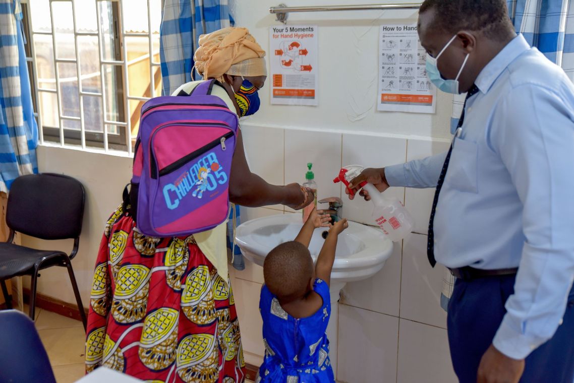 Afrikanische Frau mit Mund-Nasen-Schutz hilft einem kleinen Mädchen beim Händewaschen.
