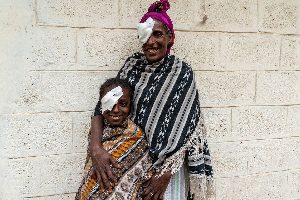 Auf dem Bild ist Mulunesh mit ihrer Mutter abgebildet. Beide tragen ein Pflaster auf dem linken Auge und lächeln.