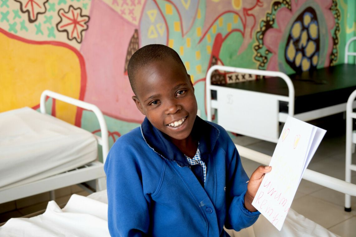 Ein lächelndes Mädchen sitzt auf einem Krankenbett. Es lächelt und hält ein Blatt Papier hoch.