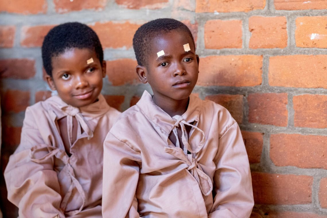 Zwei kleine Mädchen in OP-Hemden sitzen nebeneinander. Sie haben Pflaster auf der Stirn über den Augen.