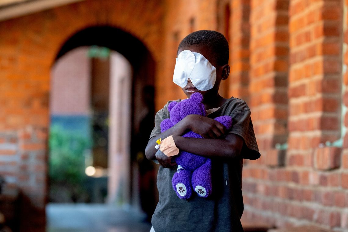 Mädchen mit Augenverbänden vor einem Backsteingebäude hält einen lila Teddybär in den Armen.