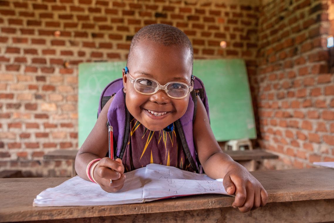 Lächelndes afrikanisches Mädchen mit Brille sitzt über ein Schulheft gebeugt.