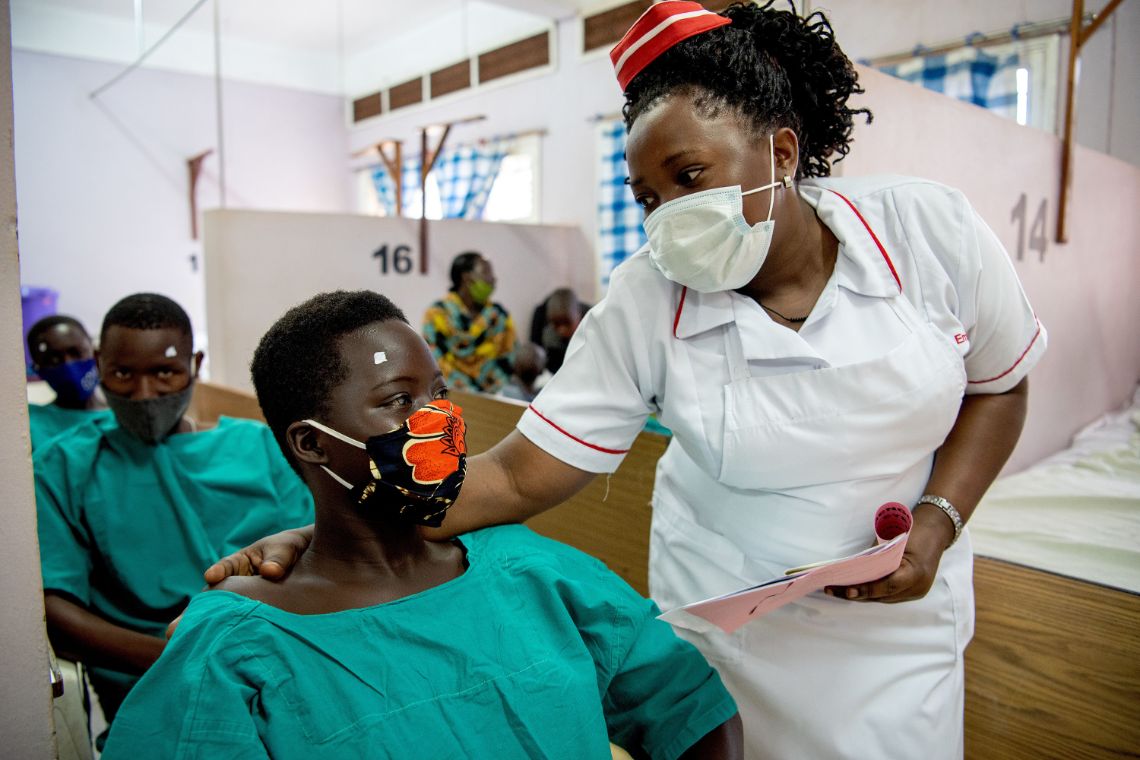 Eine Krankenschwester mit Mund-Nasen-Schutz legt einem Mädchen mit OP-Kitttel und Mund-Nasen-Schutz die Hand auf die Schulter. 