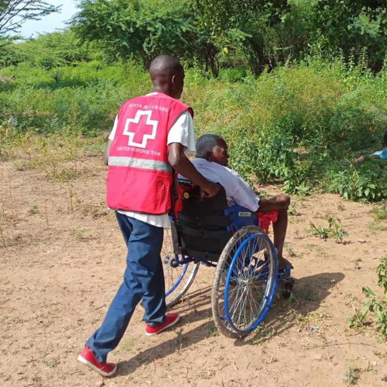 Ein Mann trägt eine Weste mit rotem Kreuz. Er schiebt einen im Rollstuhl sitzenden Mann.