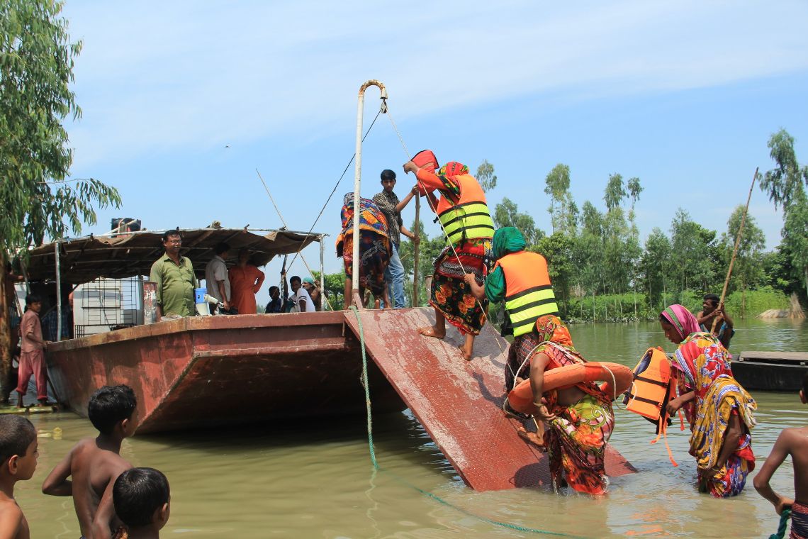 Mehre Menschen in Rettungswesten laufen aus dem Wasser über eine Rampe in ein Boot.