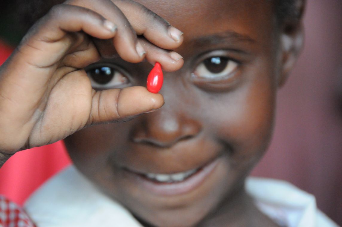 Ein afrikanisches Mädchen hält zwischen zwei Fingern eine rote Pille vors Gesicht.