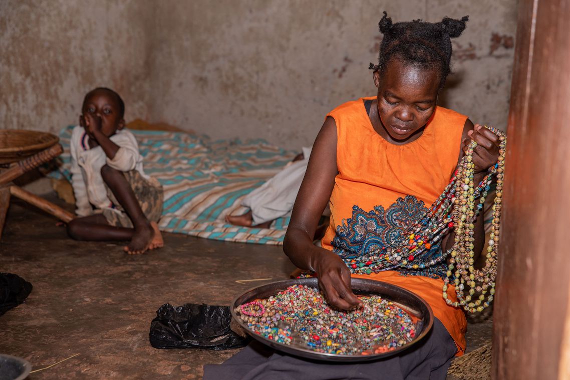 Ugandische Mutter stellt Perlenketten in ihrer Hütte her. Im Hintergrund sitzt ihr Sohn.