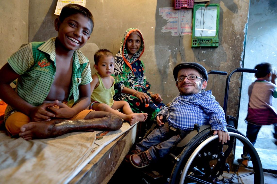 Ein Junge mit einem Bein sitzt auf einer Bank neben einem kleineren Kind und einer Frau, daneben sitzt ein Mann im Rollstuhl.