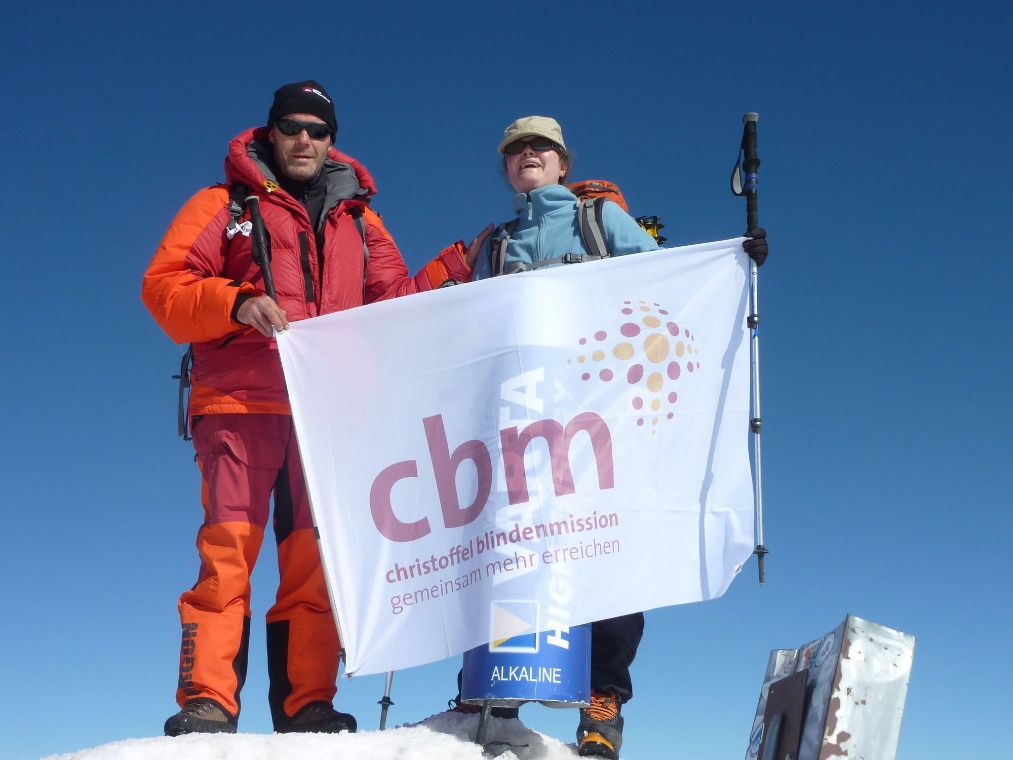 Zwei Menschen in winterlicher Bergsteigerkleidung halten eine Fahne mit CBM-Logo