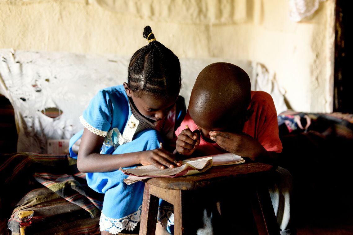 Zwei kleine afrikanische Kinder sitzen über ein Schreibheft gebeut in einer Hütte.