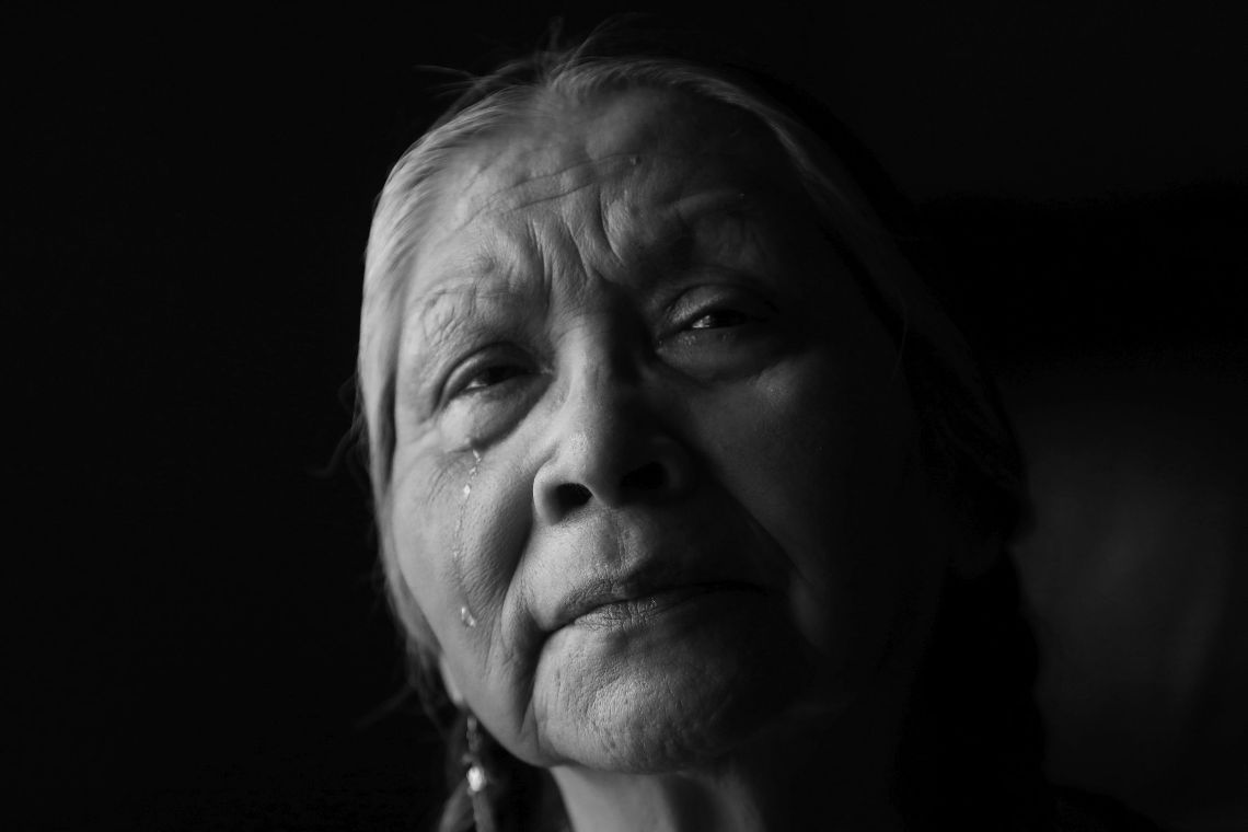 Gesicht einer älteren, indigenen Frau. Sie blickt ernst und runzelt die Stirn, eine Träne läuft ihr die Wange herunter.