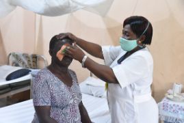 Eine Frau mit Pflaster auf dem Auge sitzt auf einem Bett. Neben ihr steht eine Krankenschwester und entfernt das Pflaster.