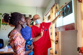 Ein kleines afrikanisches Mädchen mit extremen O-Beinen lehnt sich an einen Baumstamm.