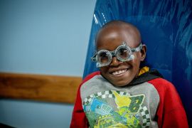 Ein kleines Mädchen ist beim Optiker auf einem Stuhl. Sie trägt eine optische Messbrille.