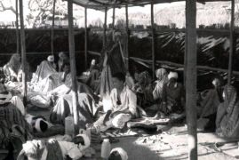 In einer Hütte aus Ästen und Planen sitzen oder liegen Männer und Frauen auf dem Boden, teils haben sie Augenverbände. Sie sind auf der Flucht vor dem Bürgerkrieg.