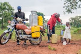 Ein mit Kanistern bepacktes Motorrad, dahinter und daneben zwei Männer und ein kleines Mädchen