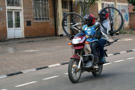 Auf der Straße fährt ein Motorradfahrer. Sein Mitfahrer auf dem Sitz hinter ihm hält mit beiden Händen ein Fahrrad fest.