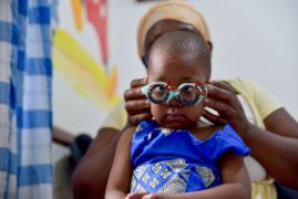 Ein afrikanischer Mann mit Mund-Nasen-Schutz hält ein Sehtestbild hoch, ein kleines Mädchen schaut darauf.