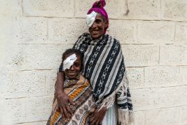 Auf dem Bild ist Mulunesh mit ihrer Mutter abgebildet. Beide tragen ein Pflaster auf dem linken Auge und lächeln.