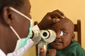 Ein kleiner afrikanischer Junge schaut in eine Lupe, die ein Mann mit Mund-Nasen-Schutz ihm vors Auge hält.