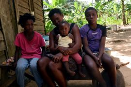 Frau, Kleinkind und zwei größere Kinder auf einer Bank vor einer Hütte