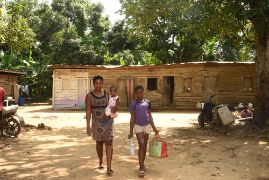 Eine Frau mit einem Kleinkind auf dem Arm und ein Junge mit einer Wasserflasche und einer Tasche auf dem unbefestigen Weg vor einem kleinen Bretterhaus.