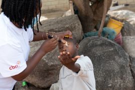 Eine Augenärztin untersucht einen Jungen am Auge. Er sitzt auf einem Stein.