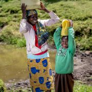 Eine Frau und ein Kind an einer Wasserstelle. Beide tragen einen Plastikkanister auf dem Kopf. Sie lachen.