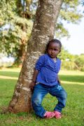 Ein kleines afrikanisches Mädchen mit extremen O-Beinen lehnt sich an einen Baumstamm.