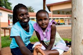 Eine afrikanische Frau und ein kleines Mädchen sitzen vor einem eingeschossigen Gebäude. Sie lachen.