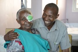 Ein afrikanischer Mann legt den Arm um eine älteres Frau mit Augenverband.