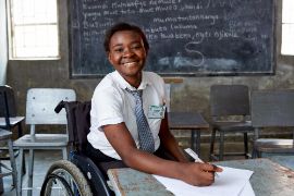 Ein afrikanisches Mädchen im Rollstuhl sitzt an einem Schulpult und lacht. 