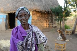 Eine afrikanische Frau mittleren Alters steht vor einer Hütte.