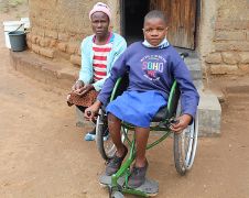 Schulmädchen in einem Rollstuhl vor einer Hütte; eine Frau sitzt daneben