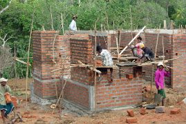 Baustelle: Männer in und vor dem Rohbau eines Backsteingebäudes, das noch kein Dach hat. Der Bau entsteht als Wiederaufbauhilfe nach einem Seebeben bzw. Tsunami.