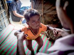 Ein Kleinkind steht auf wackeligen Beinen in einer Hütte, es wird an den Händen gehalten. Es lebt in einem Flüchtlingslager. Es gehört zur vertriebenen Minderheit der Rohingya.