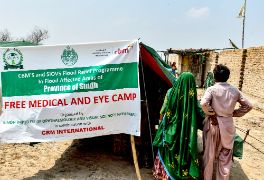 Plakat mit der Aufschrift &quot;Free Medical and Eye Camp&quot;, daneben steht eine Frau im Sari und ein Mann in Hemd und Pluderhose